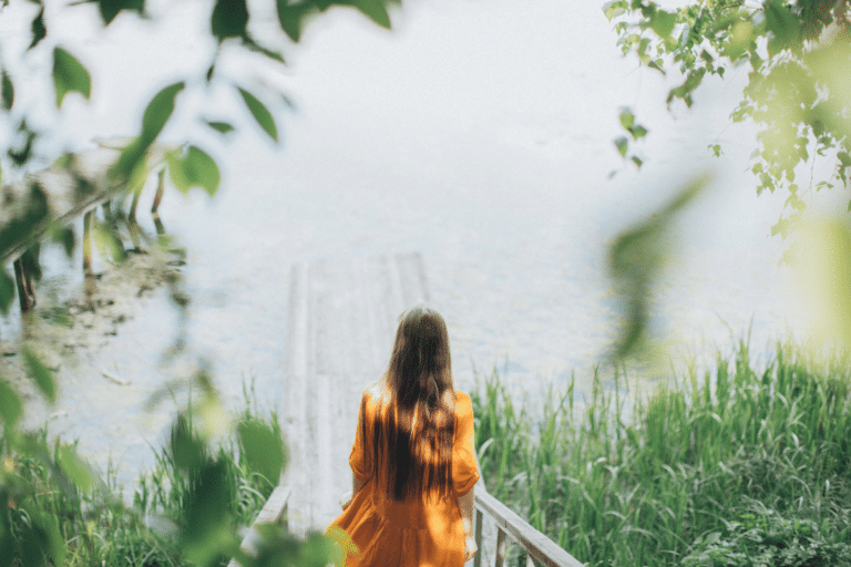 A woman in a flowing orange dress standing at the edge of a wooden dock, looking at a lake.
