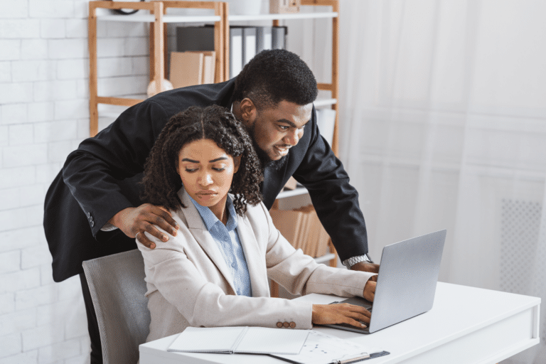 A man leaning over a seated woman at her desk, placing a hand on her shoulder, depicting workplace harassment