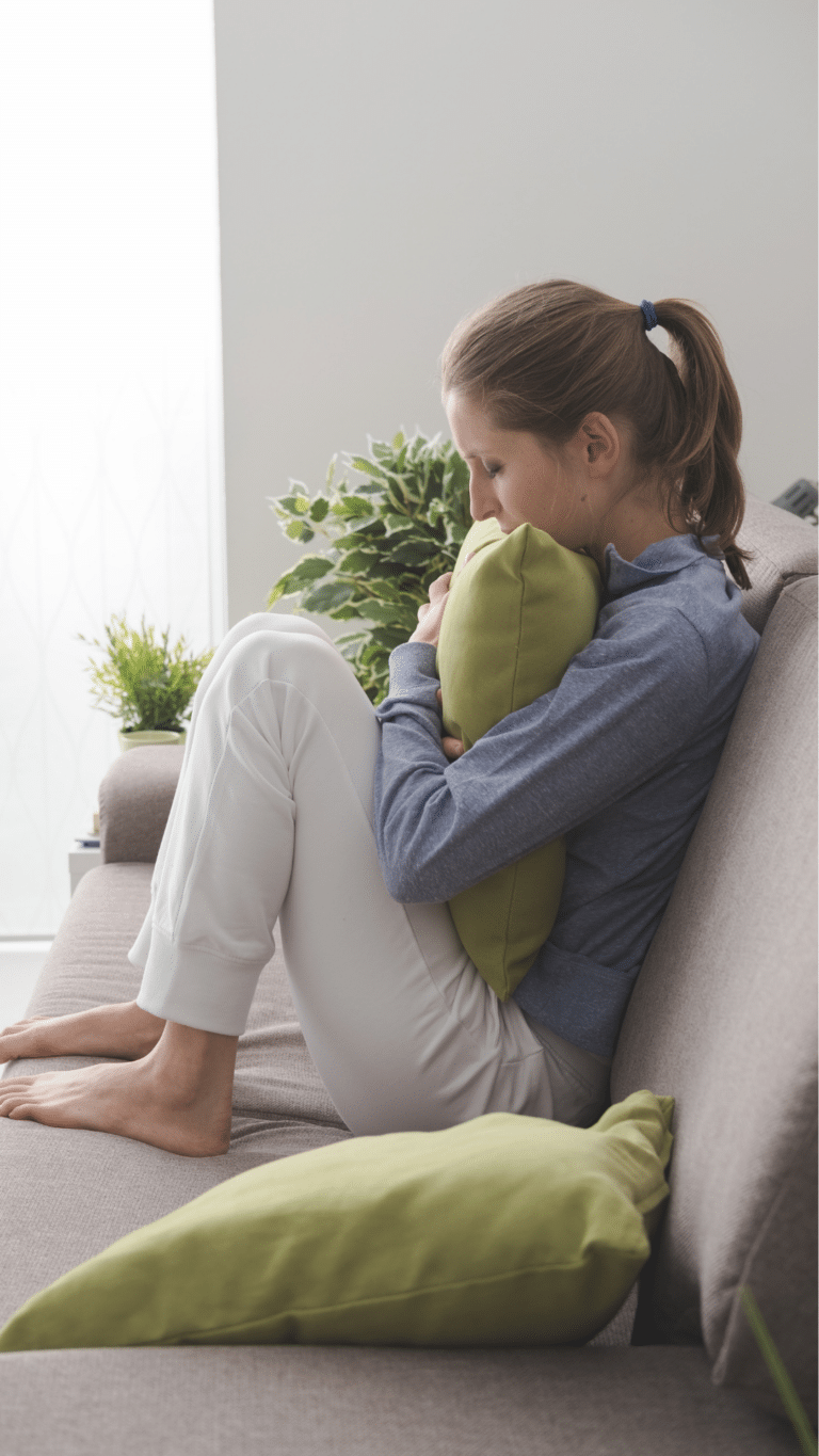A sad woman sits curled up on a couch, hugging a green pillow tightly