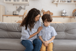 A mother sits on a couch comforting her young son, who looks upset