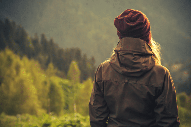 A person wearing a maroon beanie and a brown jacket stands , facing away and looking toward a lush, green forest and distant mountains