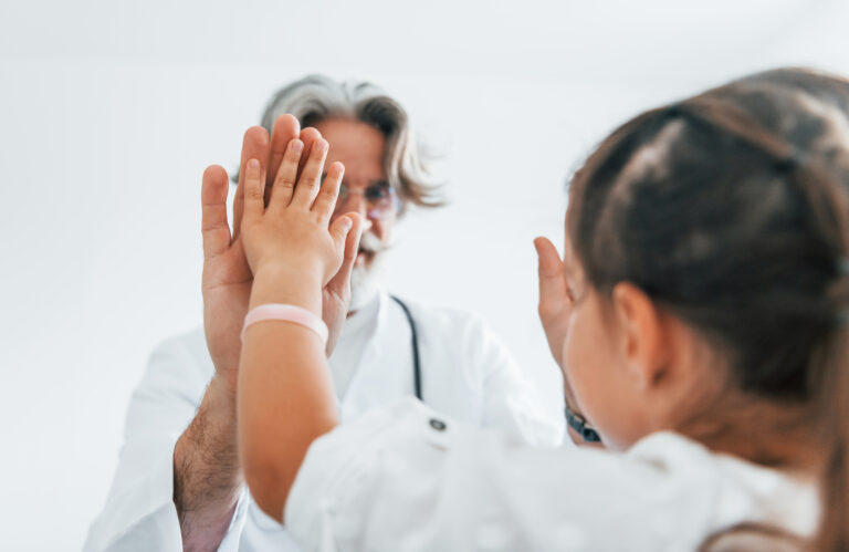 Working with little girl. Giving high five. Senior male doctor with grey hair and beard in white coat is indoors in clinic