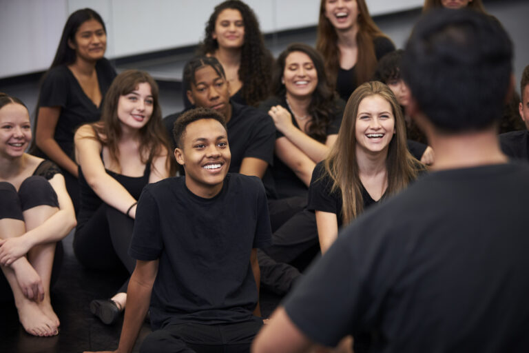 Teacher At Performing Arts School Talking To Students Sitting On Floor In Rehearsal Studio