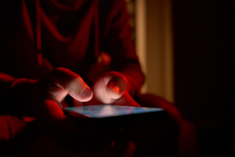 Man using smartphone at night. Close up of hand holding smartphone, swiping and scrolling social media feed on glowing screen in dark room